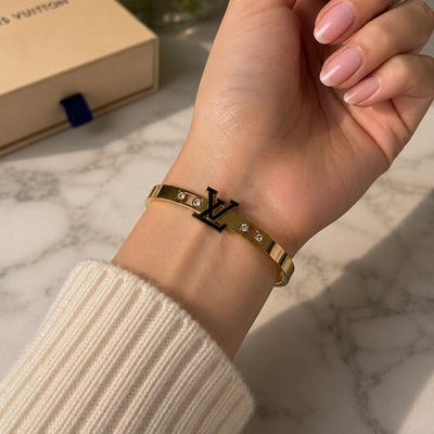 Hand wearing a gold Bangle with a Louis Vuitton logo on a marble surface with a Louis Vuitton box and flowers.