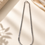 A silver-toned metal chain necklace with a curb-style link pattern, displayed in a loop on a warm cream surface with soft shadows, draped fabric, and a textured stone prop.