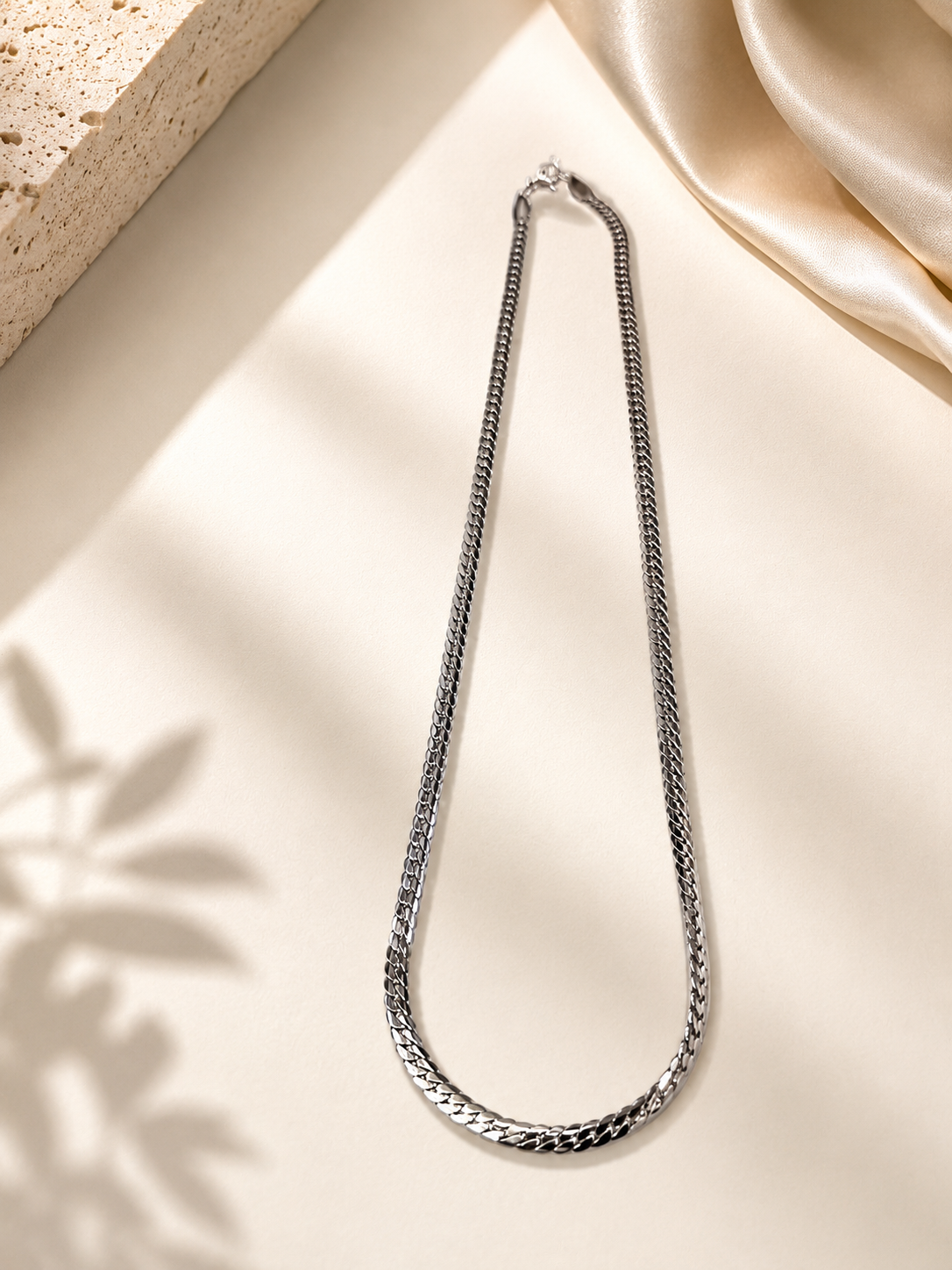 A silver-toned metal chain necklace with a curb-style link pattern, displayed in a loop on a warm cream surface with soft shadows, draped fabric, and a textured stone prop.