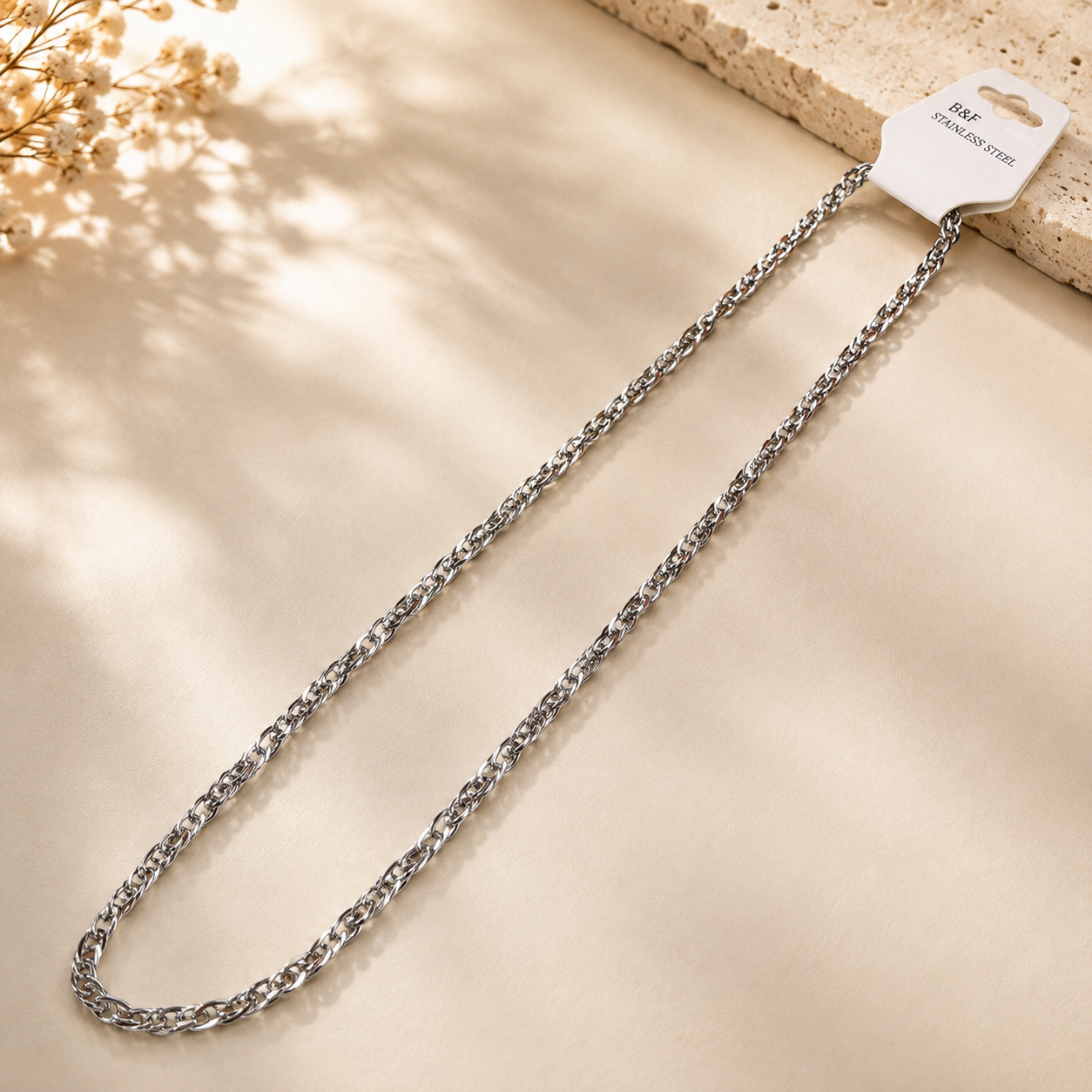 A silver-toned metal curb chain necklace with a white retail hanger tag, displayed on a textured beige surface with soft leaf shadows.