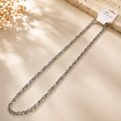 A silver-toned metal curb chain necklace with a white retail hanger tag, displayed on a textured beige surface with soft leaf shadows.
